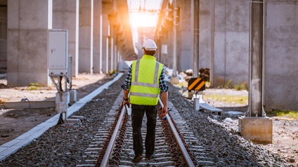 Rückenansicht einer auf Gleisen gehenden Person mit Helm, Sicherheitsweste und Tablet in der linken Hand. Links und rechts von den Gleisen sind dicke Betonpfeiler