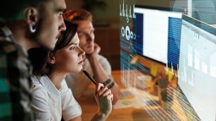 Seitliche Aufnahme einer an einem Schreibtisch sitzenden Person. Daneben stehen zwei weitere Personen, die sich am Tisch abstützen. Über dem Schreibtisch schwebt eine Projektion eines Bildschirms, auf dem Quellcodes und Statistiken sind