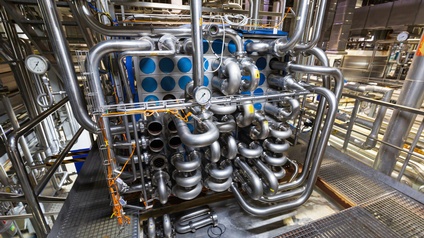 In einer Produktionsstätte stehen verschiedene große Maschinen mit Rohren und Verbindungen.