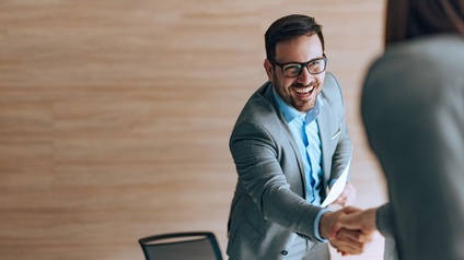 Person mit Brille und Businesskleidung gibt einer anderen Person strahlend die Hand im Stehen und hält dabei ein Blatt Papier in der Hand