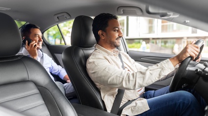 Zwei Personen in Auto sitzend, eine lenkt andere telefoniert am Rücksitz