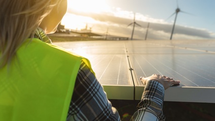 Person in Arbeitskleidung mit kariertem Hemd, gelber Warnweste und Schutzhelm blickt auf eine Photovoltaikanlage während im Hintergrund Windräder stehen, grünes Energiekonzept