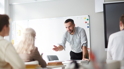 Person steht in einem Zimmer mit Whiteboard, großem Bildschirm und zeigt auf eine weitere Person, die auf einem Tisch mit Büchern sitzt während<br />
weitere Personen aufmerksam zuhören