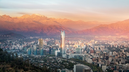 Panoramaansicht von Santiago de Chile im Dämmerlicht: Ein Wolkenkratzer ragt aus einer Stadtlandschaft vor schneebedeckten Gebirgszügen der Anden.