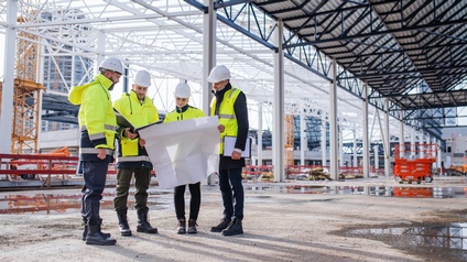 Vier Personen in gelben Sicherheitsjacken sowie mit Helmen stehen auf einer sehr großen Baustelle nebeneinander. Die zweite Person von rechts hält einen großen Bauplan. Die Person daneben hält einen aufgeklappten Ordner