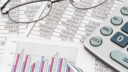 Nahaufnahme einer Brille, eines Taschenrechners und eines Stiftes, die auf bedruckten Zetteln liegen. Auf den Zetteln stehen in Tabellen verschiedene Zahlen sowie sind Statistikkurven.
