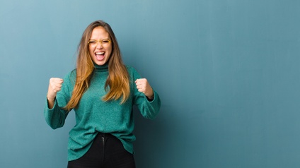 Eine Person mit dunkler Hose, grünlichem Pullover, offenen, langen Haaren sowie Brille steht vor einem grünlichen Hintergrund und ballt die Fäuste. Die Person hat den Mund geöffnet