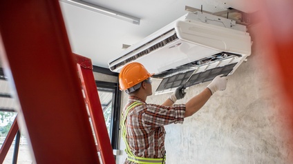 Person mit Schutzbrillen und orangem Helm hantiert an Klimaanlage