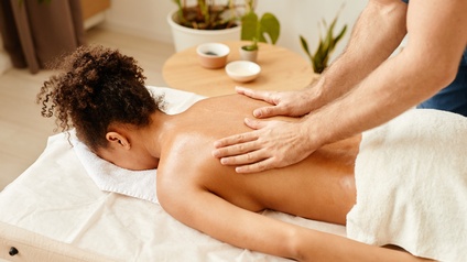 Person massiert den Rücken einer Person, die auf einer Massageliege liegt, im Hintergrund stehen Grünpflanzen
