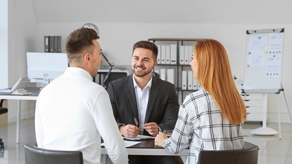 Drei Personen an Tisch in weißgrauem Büroraum sitzend im Gespräch miteinander: Zwei Personen in Rückenansicht, eine Person in Frontalansicht lächelnd, im Hintergrund verschwommen Regale mit Ordnern, Tisch mit Computer und Flipchart