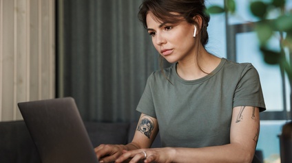 Person mit Earpods, kurzem grünen T-Shirt und Tattoos sitzt an einem Schreibtisch und tippt konzentriert in einen Laptop