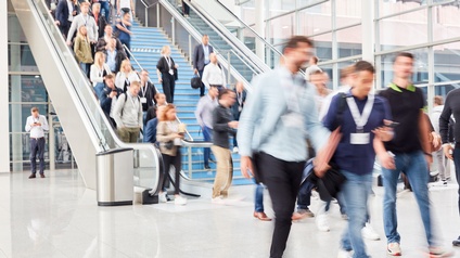 Im Vordergrund gehen Personen mit Ausweisen um den Hals. Im Hintergrund sind eine Treppe sowie eine Rolltreppe mit weiteren Personen, die hinunter und hinaufgehen