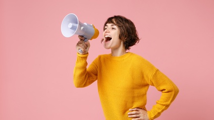 Person mit kurzen braunen Haaren, Ohrringen und gelbem Pullover hält ein Megaphon, in das sie ruft, vor einem rosa Hintergrund.