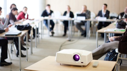 Nahaufnahme von einem Beamer in einem Besprechungsraum, im Hintergrund sitzen mehrere Personen bei Tischen 