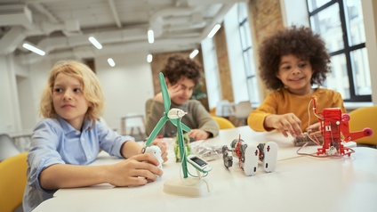 Drei Kinder sitzen an Tisch auf dem Roboter und Modell eines Windrades stehen