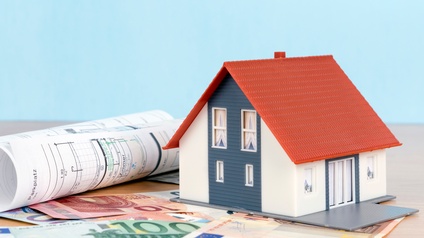 Ein Modellhaus steht auf einer hölzernen Tischplatte. Unter und vor dem Haus liegen verschiedene Eurogeldscheine, auf denen unterschiedliche Zahlen stehen. Neben dem Haus liegt eine Papierrolle, auf der Parkplatz steht. Der Hintergrund ist hellblau.