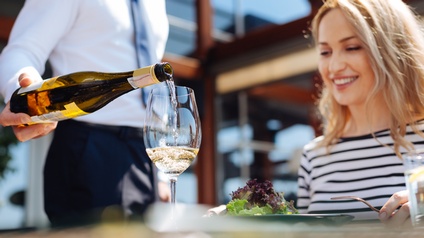 Lächelnde Person in gestreiftem Shirt sitzt vor Teller mit Salat, hält Gabel in Hand und blickt auf Glas in das andere Person aus Flasche Wein einschenkt