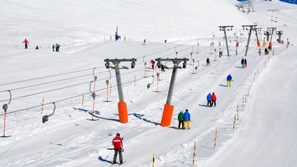 Rückenansicht von mehreren versetzen Personen au einer hügeligen, schneebedeckten Landschaft in Skikleidung, Skischuhen und Ski, die sich an schrägen Stangen festhalten, die an einem Seil befestigt sind