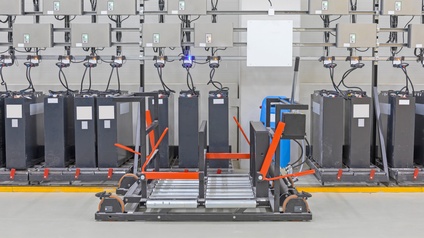 Reihe grauer Industriebatterien verbunden mit Kabeln zu grauen Boxen, im Vordergrund Hebegerät