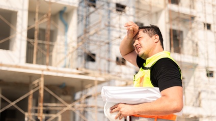 Seitliche Aufnahme einer Person mit Schutzweste, Helm und Papierrolle, die schwitzt und die rechte Hand auf die Stirn hält. Im Hintergrund ist eine Baustelle eines Gebäudes mit einem Gerüst
