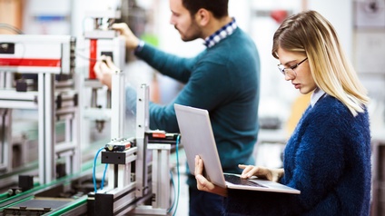 Person hält Laptop in der Hand und blickt darauf während eine weitere Person an einem technischen Objekt in einer Werkstätte arbeitet