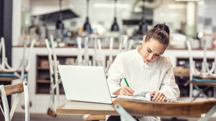 Person mit weißer Küchenarbeitskleidung sitzt an einem Tisch in einem geschlossenen Lokal indem sich die Sessel umgekehrt auf den Tischen befinden, mit ihrem Laptop und notiert sich etwas.