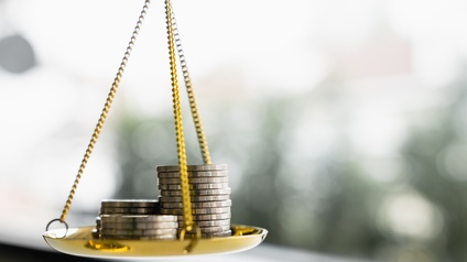 Nahaufnahme einer kleinen, goldenen Schale in der linken Bildhälfte. Am Rand sind drei goldene, kleine Ketten montiert, die oben aufeinander treffen. Auf der Schale liegen drei Münzstapel