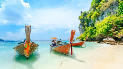 Im türkisblauen Meer stehen drei kleinere Boote nebeneinander. Daneben ist weißer Sandstrand. Neben dem Meer ist ein steiniger Hügel mit grünen Bäumen