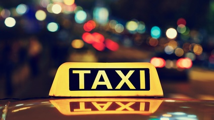 Leuchtend gelbes Zeichen auf Autodach: Gelber länglicher Leuchtkörper mit schwarzen Elementen und Schriftzug Taxi spiegelt sich in Autodach, im Hintergrund verschwommen bunte Lichtreflektionen in der Dunkelheit