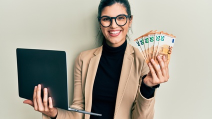 Halbporträt einer lächelnden Person in einem schwarzen Oberteil mit Sakko und Brillen, die vor einer hellen Wand steht. In der rechten Hand hält sie einen aufgeklappten Laptop. In der linken Hand hält sie mehrere Fünfzig-Eurogeldscheine.