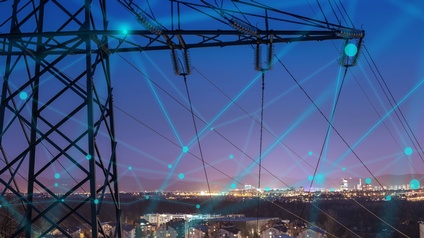 Im Vordergrund im Ausschnitt Strommasten mit blauem Overlay zu Punkten zusammenlaufender Linien ein Netz formend, im Hintergrund Stadtlandschaft im Abendlicht