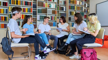 Eine Gruppe von Schüler:innen sitzt in einem Halbkreis in einer Bibliothek mit einer Lehrperson und spricht miteinander.