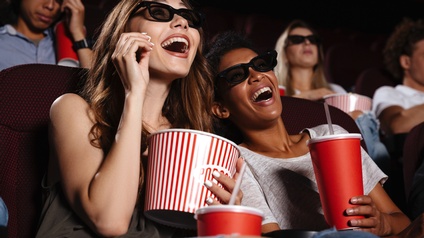 Portrait von zwei Personen, die lachend mit einer 3D-Brille, Popcorn und Softdrinks in einem Kinosaal sitzen, daneben und dahinter sitzen weitere Personen