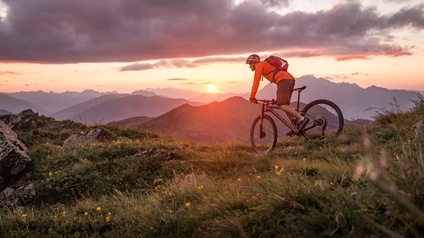 Person mit Helm und Rucksack auf Mountainbike fährt auf Wiese, im Hintergrund Berglandschaft im Sonnenuntergang