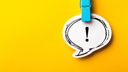 Auf gelblichem Hintergrund ist in der rechten Bildhälfte eine aus Papier ausgeschnittene Sprechblase, in der ein Rufzeichen ist. Auf der Sprechblase oben ist eine türkise Wäscheklammer aus Holz