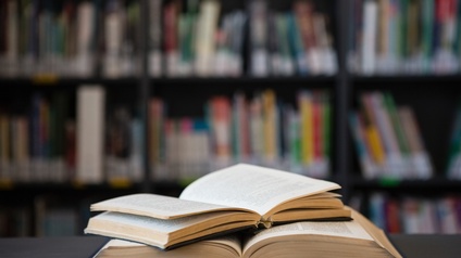 Zwei aufgeklappte Bücher liegen aufeinander, im Hintergrund verschwommen stehen volle Bücherregale.