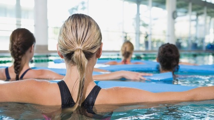 Rückenansicht der Köpfe von vier Personen. Die Personen stehen in einem Wasserbecken. Vor jeder Person ist ein längliches, blaues, schwimmendes Objekt. Die Personen haben ihre Hände darauf liegen.