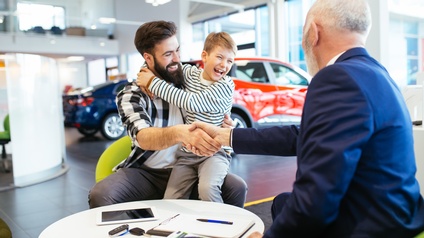 Person mit lachendem Kind am Schoß schüttelt weiterer Person am Tisch sitzend die Hand, im Hintergrund Autos