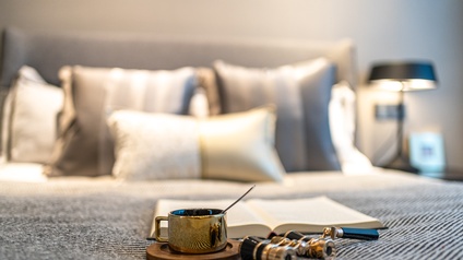 Auf der Decke eines gemachten Bettes sind eine Tasse, ein Operngucker und ein aufgeklapptes Buch. Im Hintergrund sind mehrere Pölster sowie ein Nachtkästchen mit einer Stehlampe