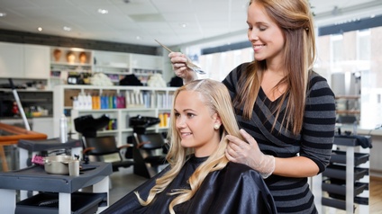 Lächelnde Person mit Plastikhandschuhen hält Pinsel mit Paste an Haarscheitel anderer vor ihr sitzender Person mit langen blonden Haaren, im Hintergrund verschwommen Friseursessel mit Kopfwaschbecken und diverse Friseurutensieien