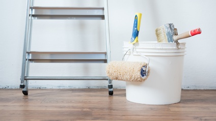 An einer weißen Wand ist eine Leiter angelehnt. Daneben steht ein weißer Eimer, in dem ein Malerpinsel und eine Farbrolle sind.