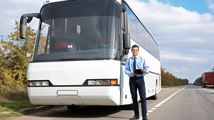 Ein weißer Autobus parkt neben einer Fahrspur. Vor dem Reisebus steht eine Person, die ein Tablet hält. Auf den Fahrspuren fahren Autos. Links neben dem Bus sind grüne Büsche