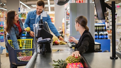 Zwei Personen legen auf das Förderband einer Kassa in einem Supermarkt verschiedene Lebensmittel wie Äpfel aus einem Einkaufswagen. Eine weitere Person sitzt hinter der Kassa