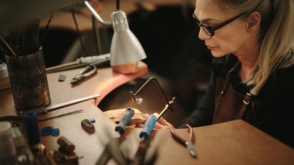 Seitliche Aufnahme einer an einer erhöhten Holzplatte sitzenden Person. An der Platte ist ein kleines unebenes Holzstück angebracht, an das die Person eine kleine Säge hält. Auf der Platte liegen weitere Werkzeuge wie Zangen