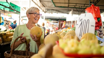 Seitliche Aufnahme einer Person, die in einem Supermarkt vor verschiedenen Obstsorten wie Bananen oder Mandarinen steht und in der linken Hand einen Granatapfel hält. Um die linke Schulter hat sie einen Einkaufskorb