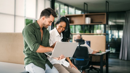 Zwei Personen sitzen nebeneinander auf einem Tisch in einem Büro. Die linke Person hält einen aufgeklappten Laptop. Die rechte Person blickt auf den Bildschirm des Laptops. Im Hintergrund sitzt eine Person an einem Schreibtisch