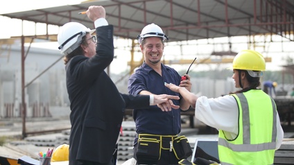 Drei Personen mit Schutzhelmen in Baustellenumgebung schlagen jubelnd die Hände aufeinander
