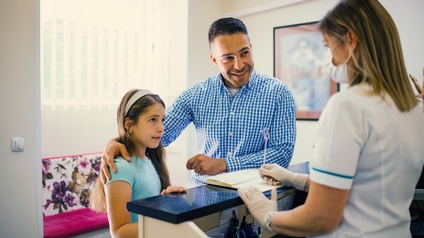 Erwachsene Person und Kind stehen an Rezeption einer medizinischen Ordination, Person hinter Rezeption trägt Mundnasenschutz und Gummihandschuhe und nimmt Notizen in Kalender vor