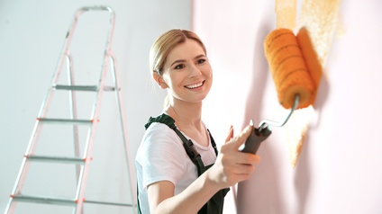 Lächelnde Person in Berufskleidung malt mit Malerrolle orange Farbe auf Wand, im Hintergrund steht eine Leiter