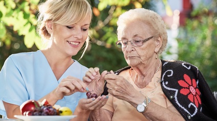 Pflegekraft unterstützt ältere, betagte Person mit Brille beim Schälen von Obst